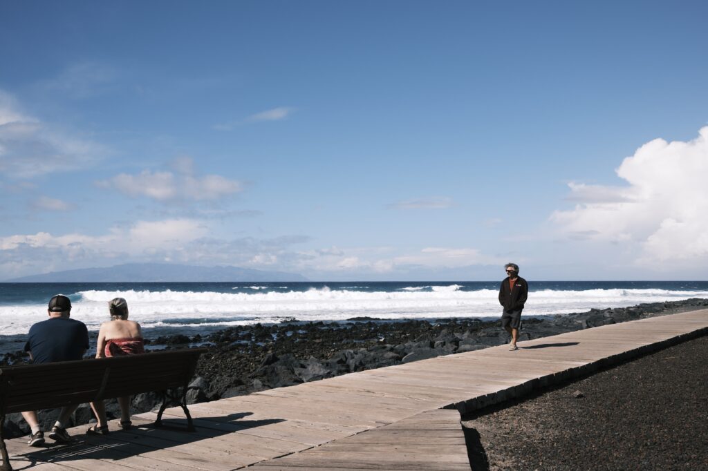 Ein Paar sitzt auf einer Bank am Holzsteg und schaut aufs Meer, während ein Mann an der Küste entlang spaziert, mit Wellen im Hintergrund.