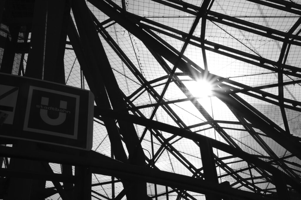 Schwarz-Weiß-Aufnahme einer komplexen Stahlkonstruktion mit Sonnenstrahlen und einem Schild für die Stadtbahn (U-Bahn).