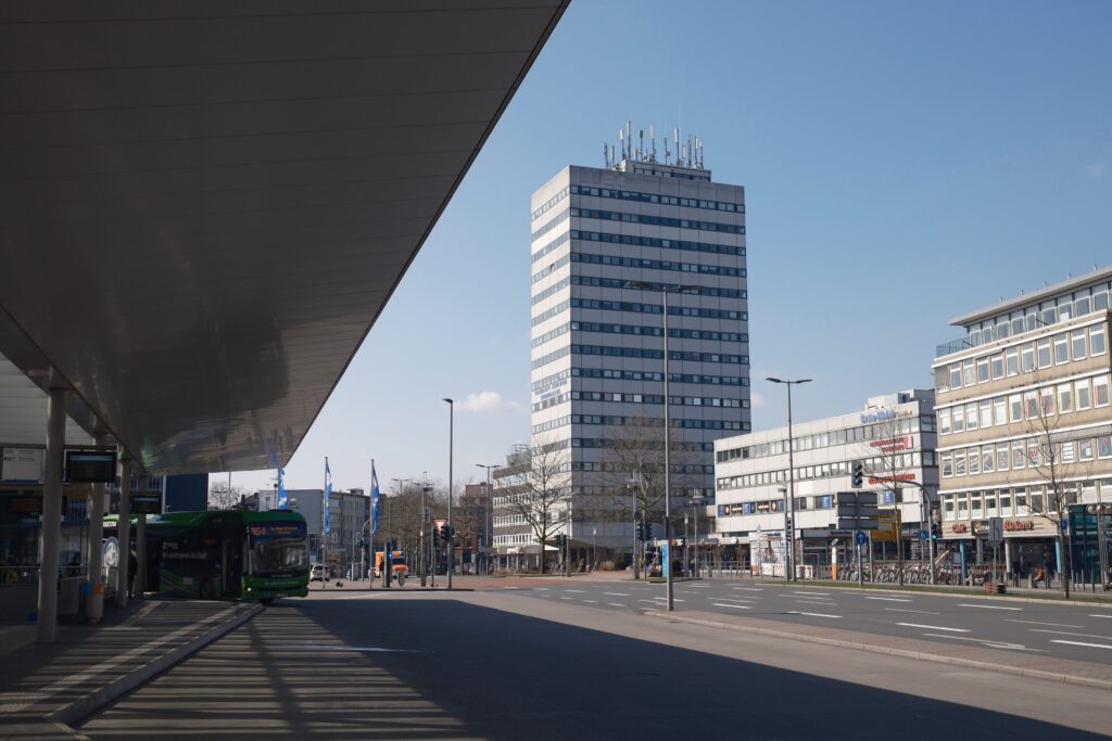 Blick auf eine städtische Szenerie mit einem Busbahnhof, einem grünen Linienbus und einem hohen Bürogebäude im Hintergrund bei klarem Himmel.