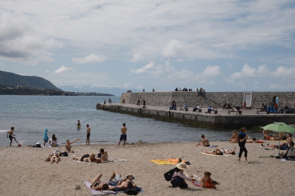 Menschen entspannen am Strand von Cefalu, im Hintergrund die markante Mole und das Meer unter leicht bewölktem Himmel