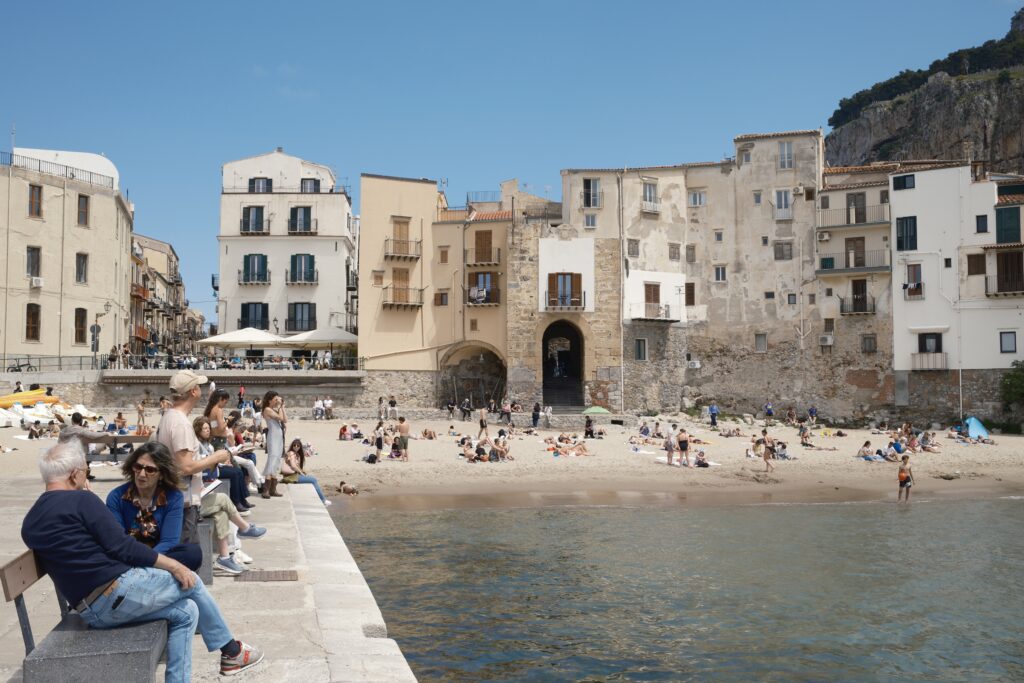 Blick vom Wasser auf die Altstadt von Cefalu mit Strand, Badenden und historischem Stadttor