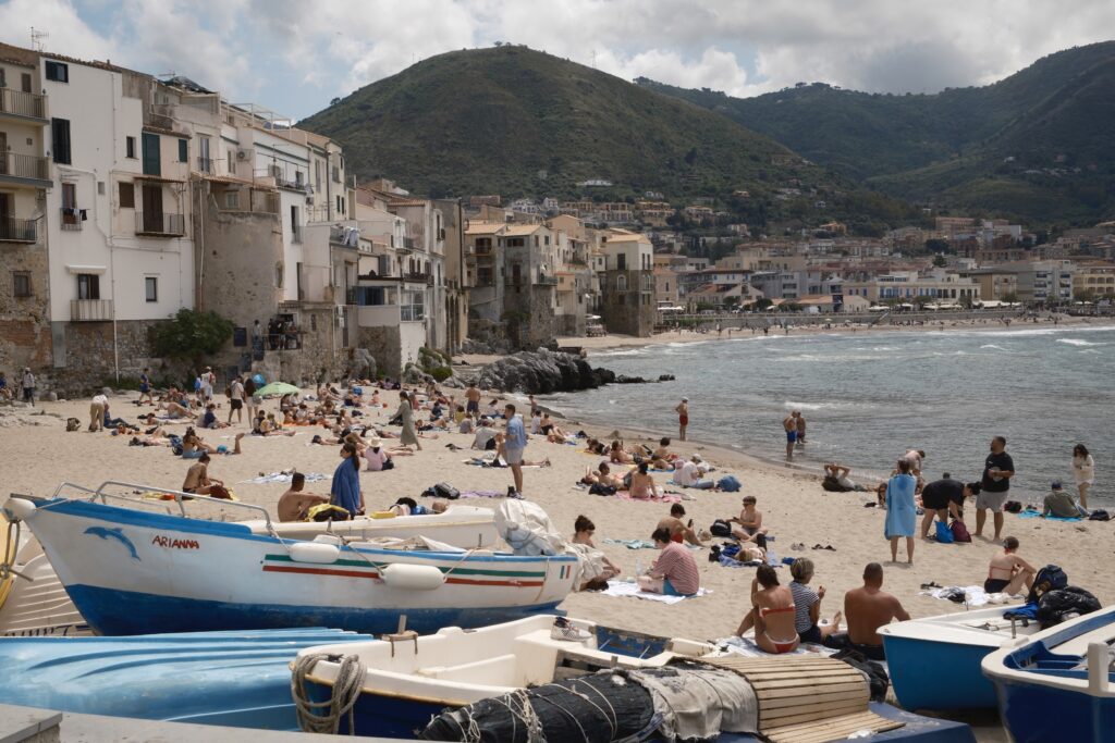 Fischerboote am Strand von Cefalu, dahinter Badende, die Mole und die grünen Berge der sizilianischen Küste
