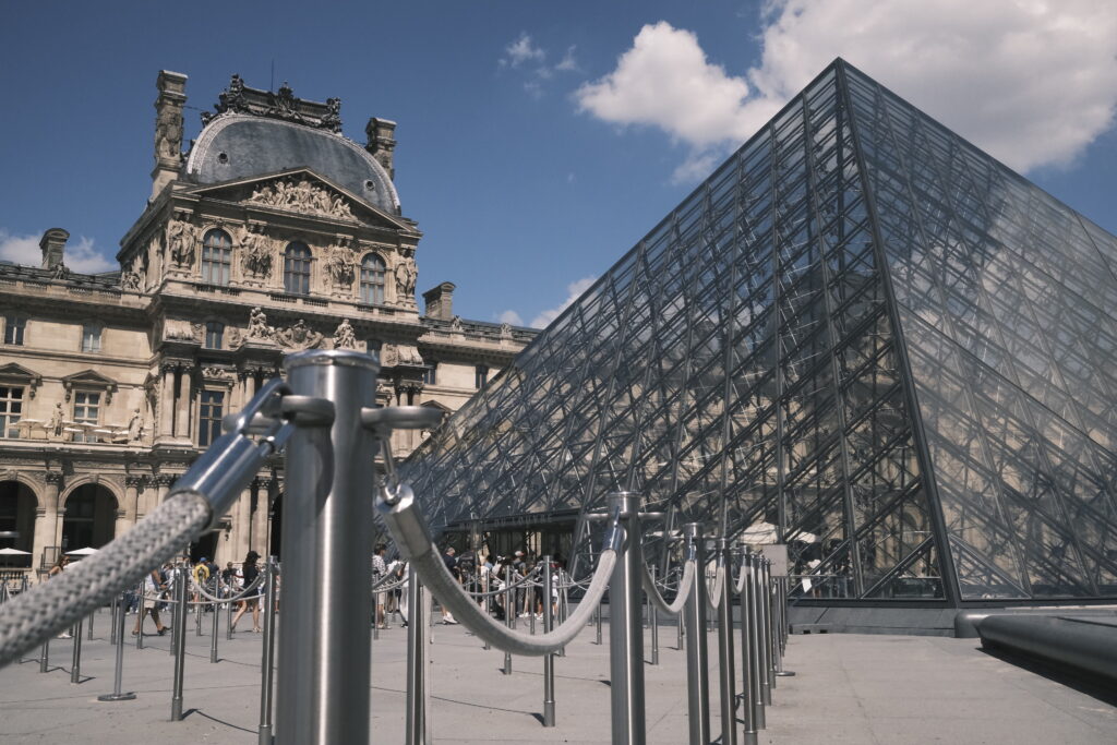 Die Glaspyramide des Louvre-Museums in Paris im Sonnenlicht, davor historische Museumsfassade.