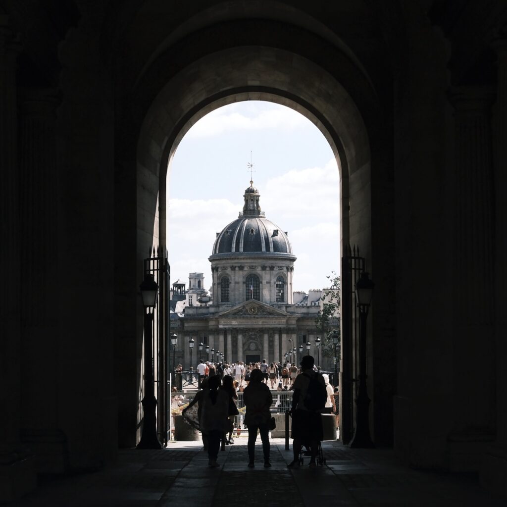 Blick durch einen Torbogen auf das Institut de France bei Tageslicht, Silhouetten von Passanten im Vordergrund.