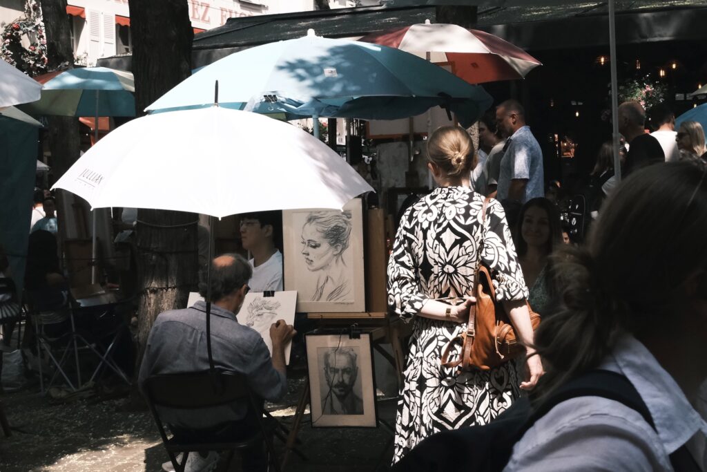 Straßenszene am Place du Tertre mit mehreren Künstlern unter Sonnenschirmen, die Skizzen und Malereien präsentieren.