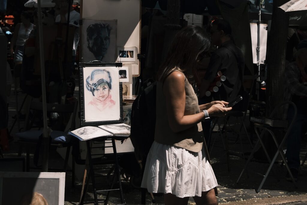 Eine junge Frau, die an einem der belebten Stände mit Porträts vorbeigeht. Im Hintergrund reihen sich Zeichnungen und Skizzen aneinander.