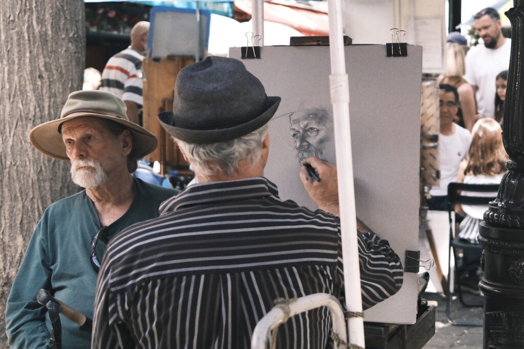 Straßenkünstler zeichnet ein Porträt auf einem Marktplatz in Paris, ein älterer Mann sitzt Modell.