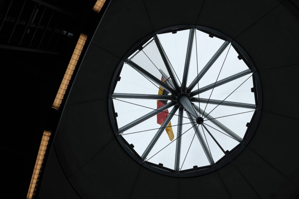 Blick von innen durch eine runde Deckenöffnung auf die wehende deutsche Flagge und moderne Dachkonstruktion des Bundestags.