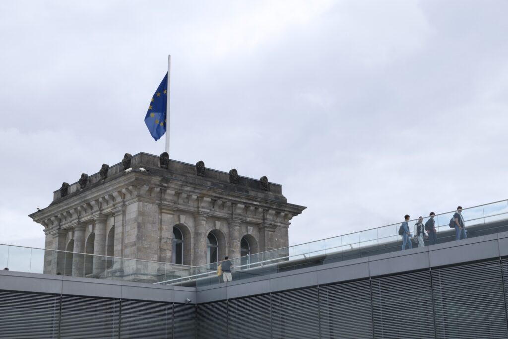 Historischer Eckturm des Berliner Reichstags mit europäischer Flagge, im Vordergrund eine moderne Glasbrüstung und Besucher.