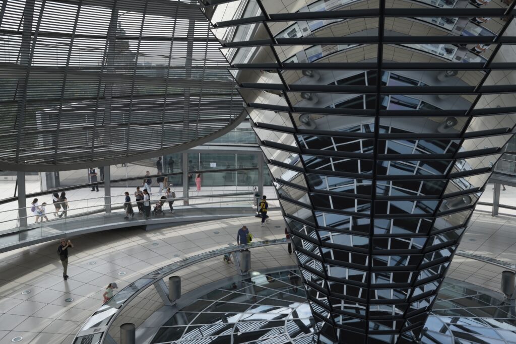 Innenaufnahme der Reichstagskuppel mit zentralem Spiegelkegel, Reflektionen, laufenden Besuchern und transparenter Architektur.