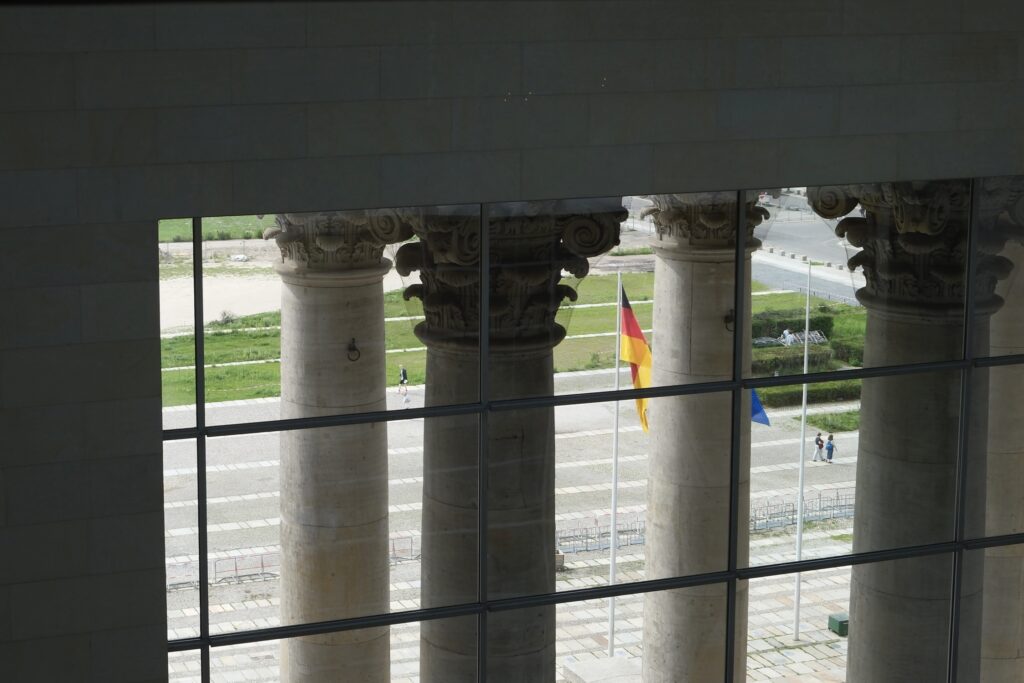 Blick durch ein großes Fenster auf historische Säulen des Reichstags, davor deutsche und europäische Flagge sowie Spaziergänger.
